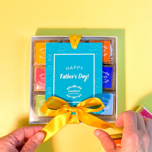 Box with clear top showing brownies with colourful labels inside. on the top of the box there is a blue tag written Happy Father's Day and yellow ribbon with two hands open the ribbon.