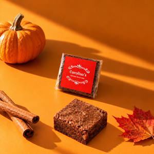 brownie with red label and one unwrapped brownie on a yellow table next to cinnamon sticks and maple leaves, in the background there is a small pumpkin
