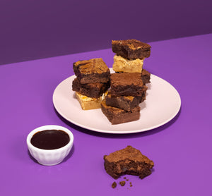 Brownies on a white plate with a small bowl of chocolate sauce on a purple background