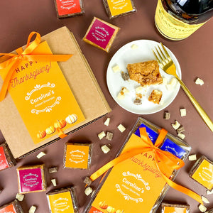 Gift box with thanksgiving packaging and brownies scattered around them on the table. a bottle of wine on the top right and a plate with a brownie and a golden fork in the centre.