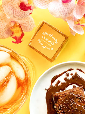 brownie wrapped with yellow label on a yellow background in the middle of orchids on top, a brownie with chocolate sprinkles and some sauce on a white plate and a glass of iced tea.