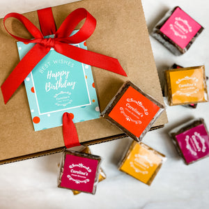 A box of gourmet brownies with a 'Happy Birthday' card and red ribbon, with several individually wrapped brownies displayed in front.