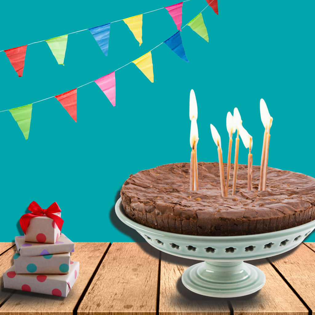 Chocolate cake with lit candles on a white cake stand, surrounded by colorful flags and gift boxes.