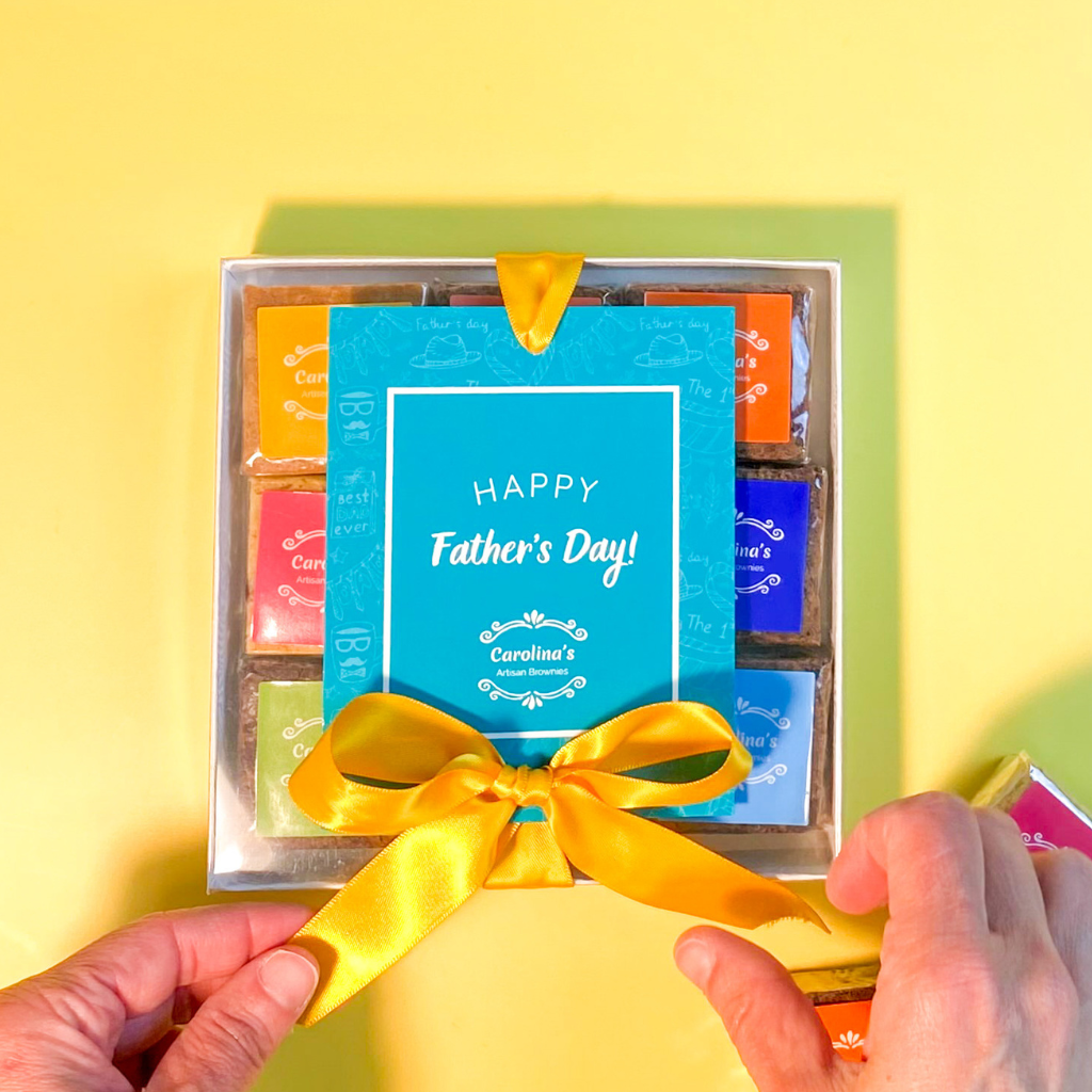 Box with clear top showing brownies with colourful labels inside. on the top of the box there is a blue tag written Happy Father's Day and yellow ribbon with two hands open the ribbon.