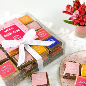 Carolina’s Gift Box of gourmet brownies with 32 brownies and hand picking up a brownie from a pink plate. Vase in the background 