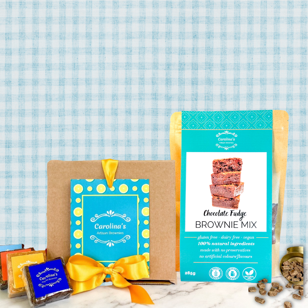 A gift set featuring a 300g bag of Carolina's Chocolate Brownie Mix, a brown kraft gift box with a blue and yellow card, several individually wrapped chocolates, and a yellow ribbon, arranged on a marble surface against a blue and white gingham background.