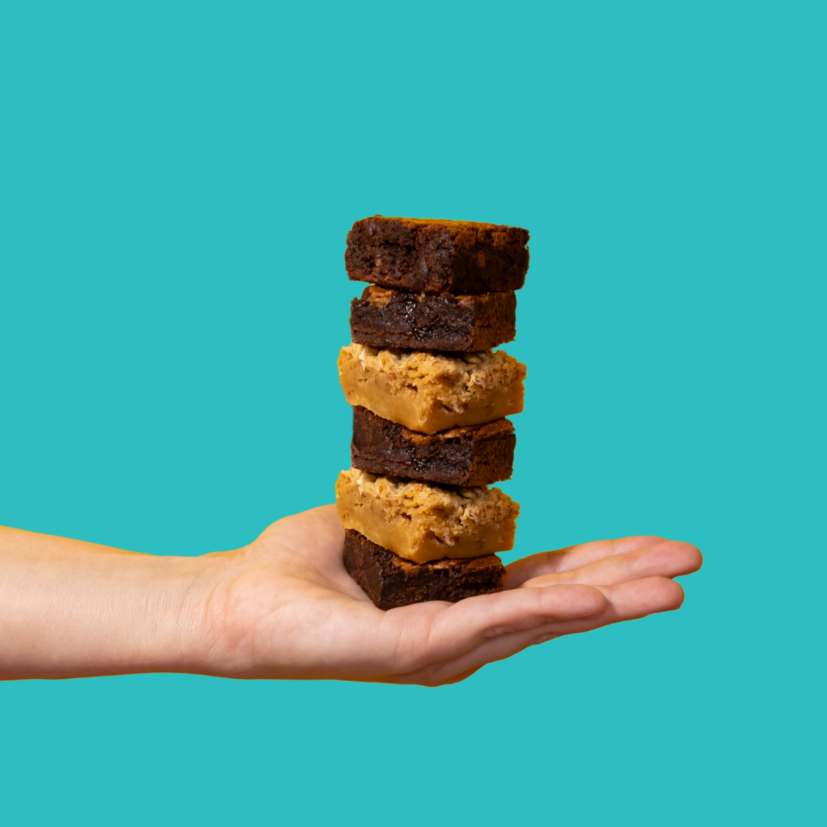 A hand holding a stack of six brownies with a variety of toppings and fillings, on a teal background.