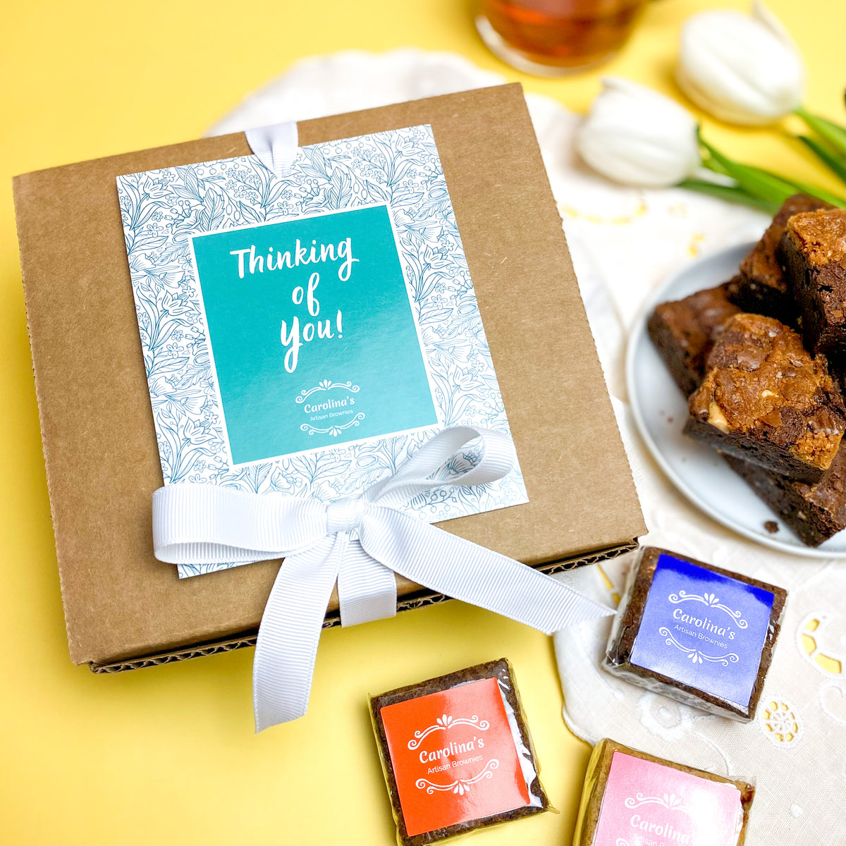A gift box of handmade brownies with a 'Thinking of You' card, presented on a yellow background with a plate of brownies and decorative tulips.