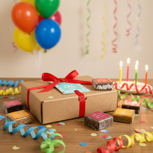 box of Carolina's brownies on a birthday table with scattered brownies and birthday decorations. candles and balloons in the background.