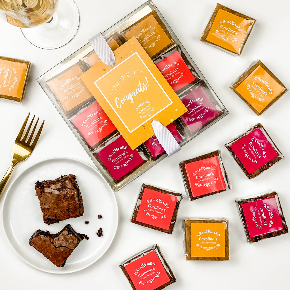 A selection of gourmet brownies in various colors and flavors, packaged in a clear box with a 'Congrats' card, alongside a piece of chocolate brownie on a plate with a fork.