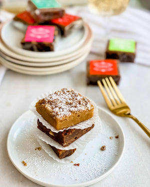 pile of three brownies on a white plate with scattered brownies on the back and a golden fork on the right side