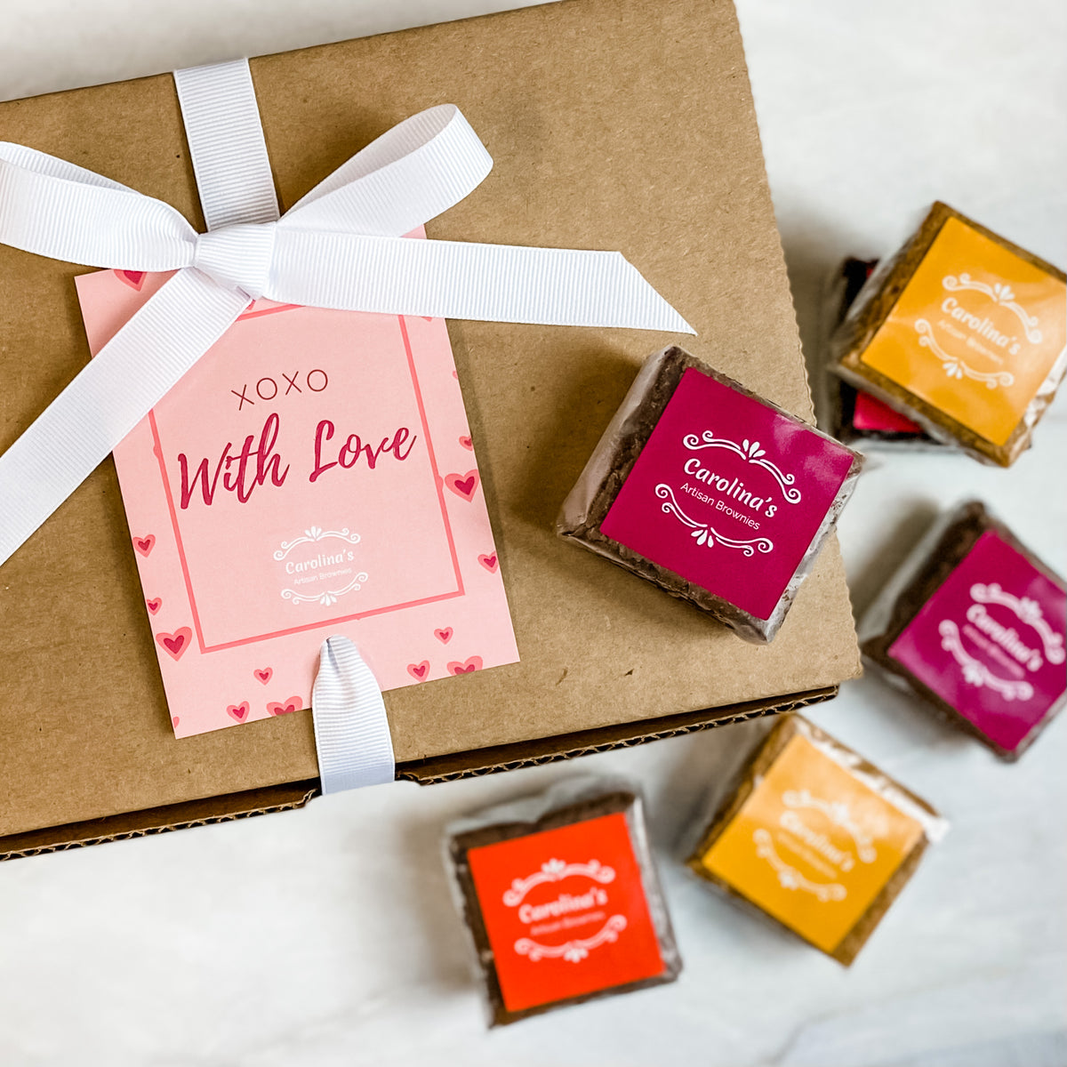 A gift box with a pink 'XOXO With Love' tag, containing assorted gourmet brownies wrapped in individual packaging with ribbon.
